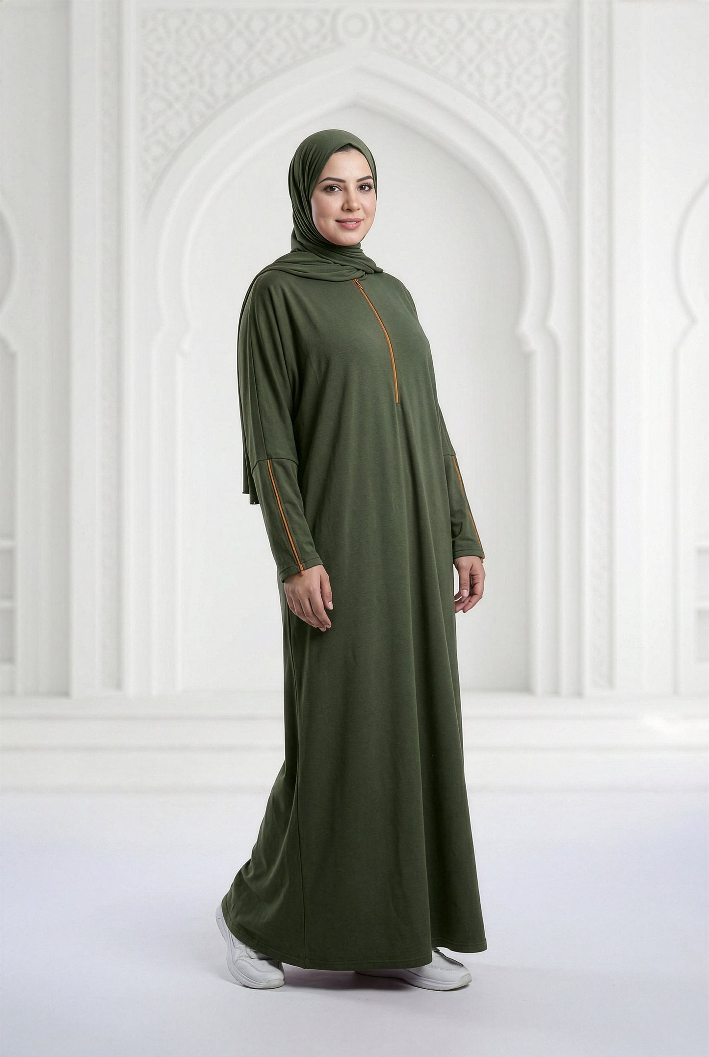 Woman wearing a dark olive Oumra Isdal with embroidered front and sleeve detailing, modest flowing silhouette, standing in an elegant indoor archway