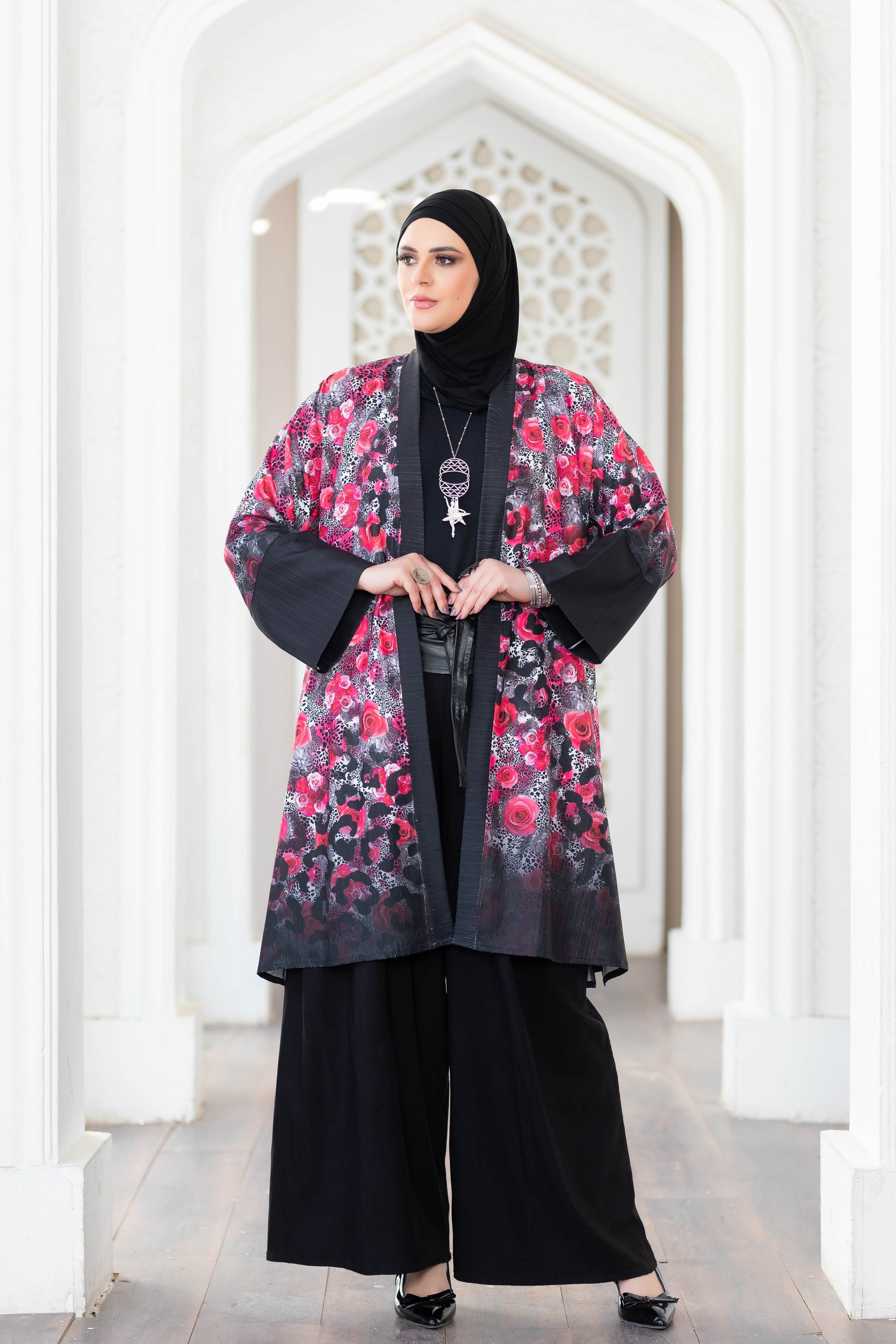 Colorful floral print kimono with black sleeves and panels, worn over a black outfit by a woman in a traditional headscarf, standing in an architectural white arched doorway.