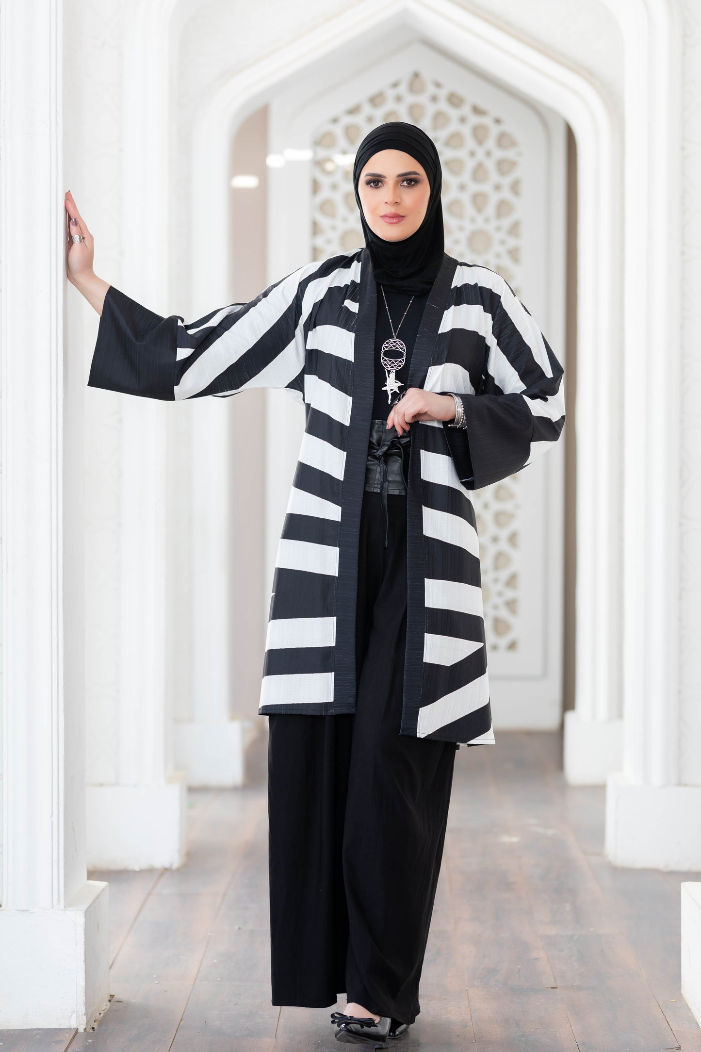 Stylish black and white striped kimono with flowing sleeves, worn by a woman in a modest Islamic outfit, standing in an archway with a floral pattern.