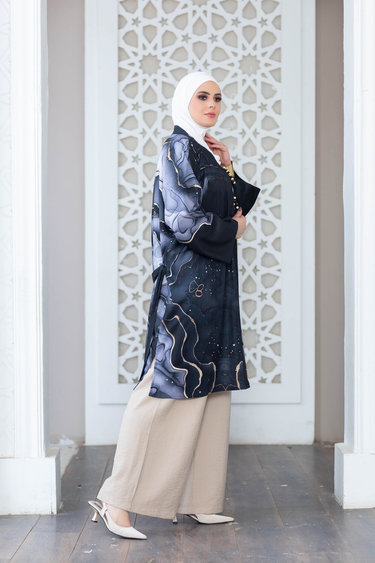 Stylish modern Islamic attire: patterned kimono, long flowing dress, and modest headscarf. The woman stands confidently in front of an intricate architectural backdrop.