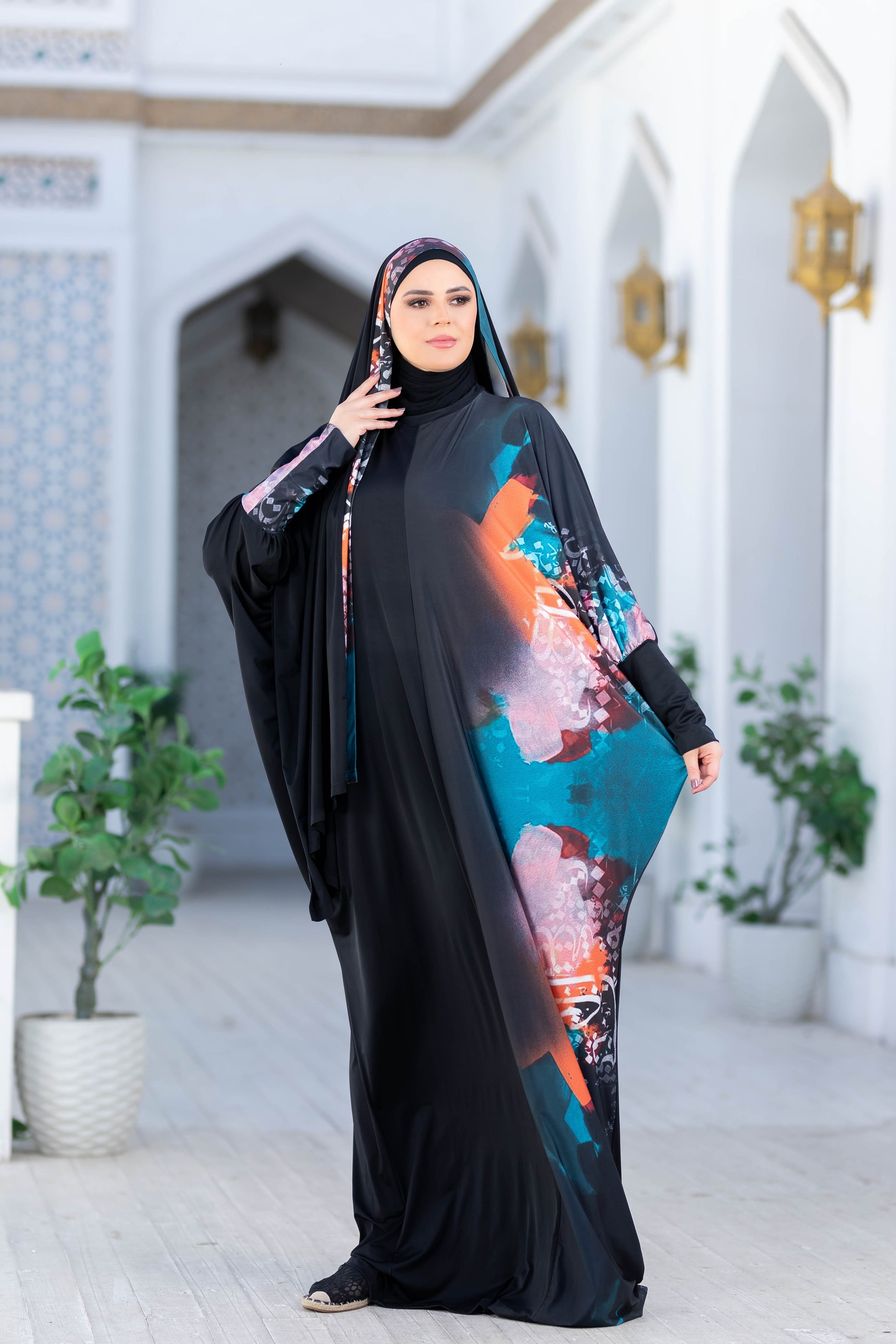 Modest Isdal 777179 ESDAL - Elegant black abaya with vibrant floral print accents, worn by a woman indoors with traditional Middle Eastern decor.