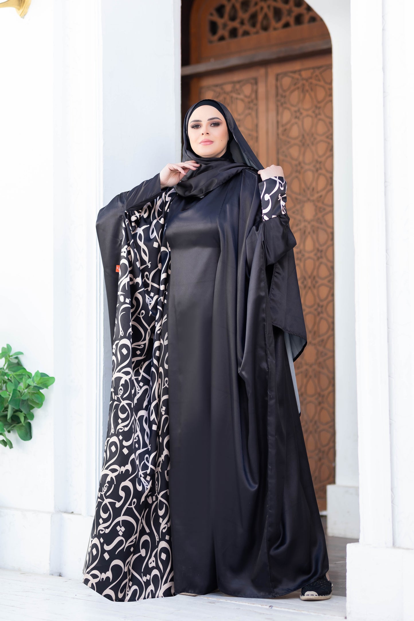 Modest satin Isdal 777189 abaya with intricate floral pattern, worn by a woman against an ornate architectural backdrop.
