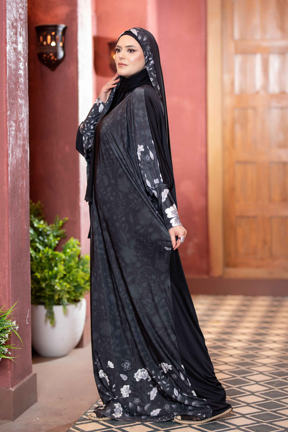 Elegant Islamic attire: Black floral-patterned modest dress with long sleeves, paired with a hijab, showcased against a traditional architectural background.