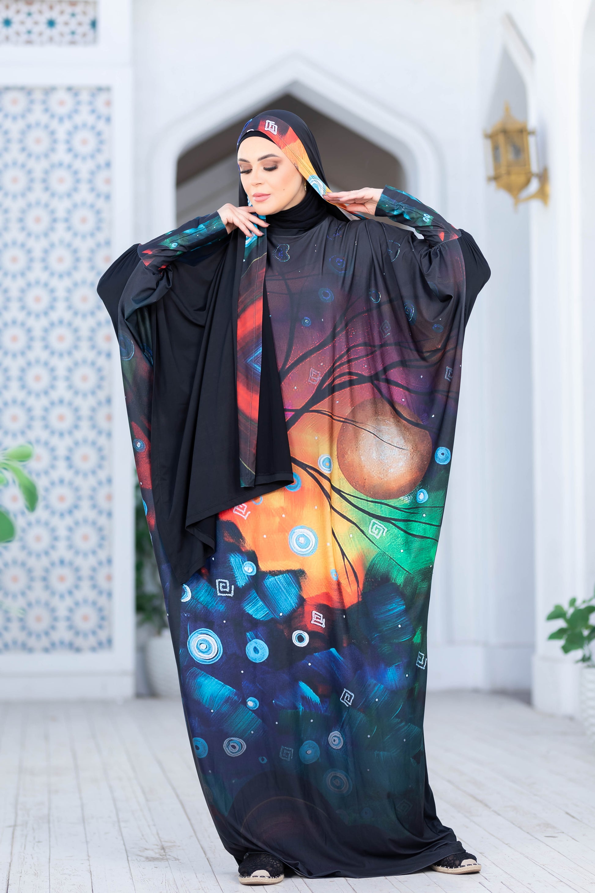Vibrant abstract print modest fashion dress, featuring colorful circles and patterns on a dark background. The dress is worn by a woman in a traditional headscarf, posing in an indoor setting with decorative architectural elements in the background.