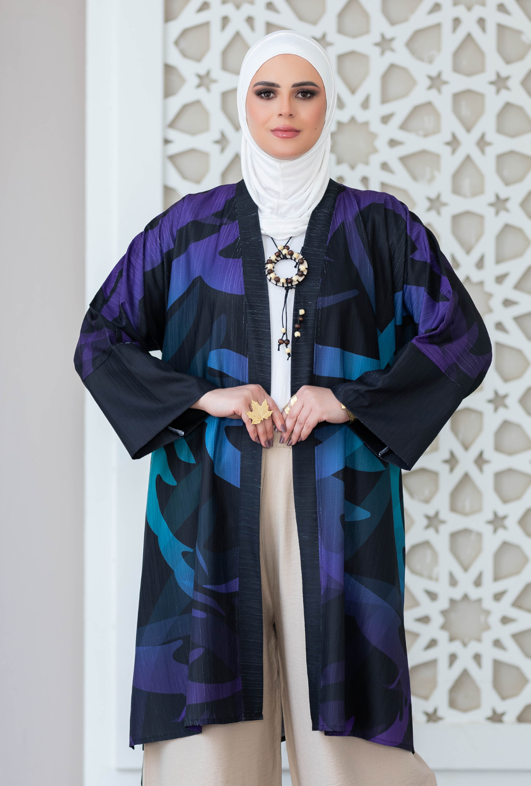 Stylish patterned kimono jacket worn with hijab, pendant necklace, and neutral pants by a young woman posing in front of a decorative architectural backdrop.