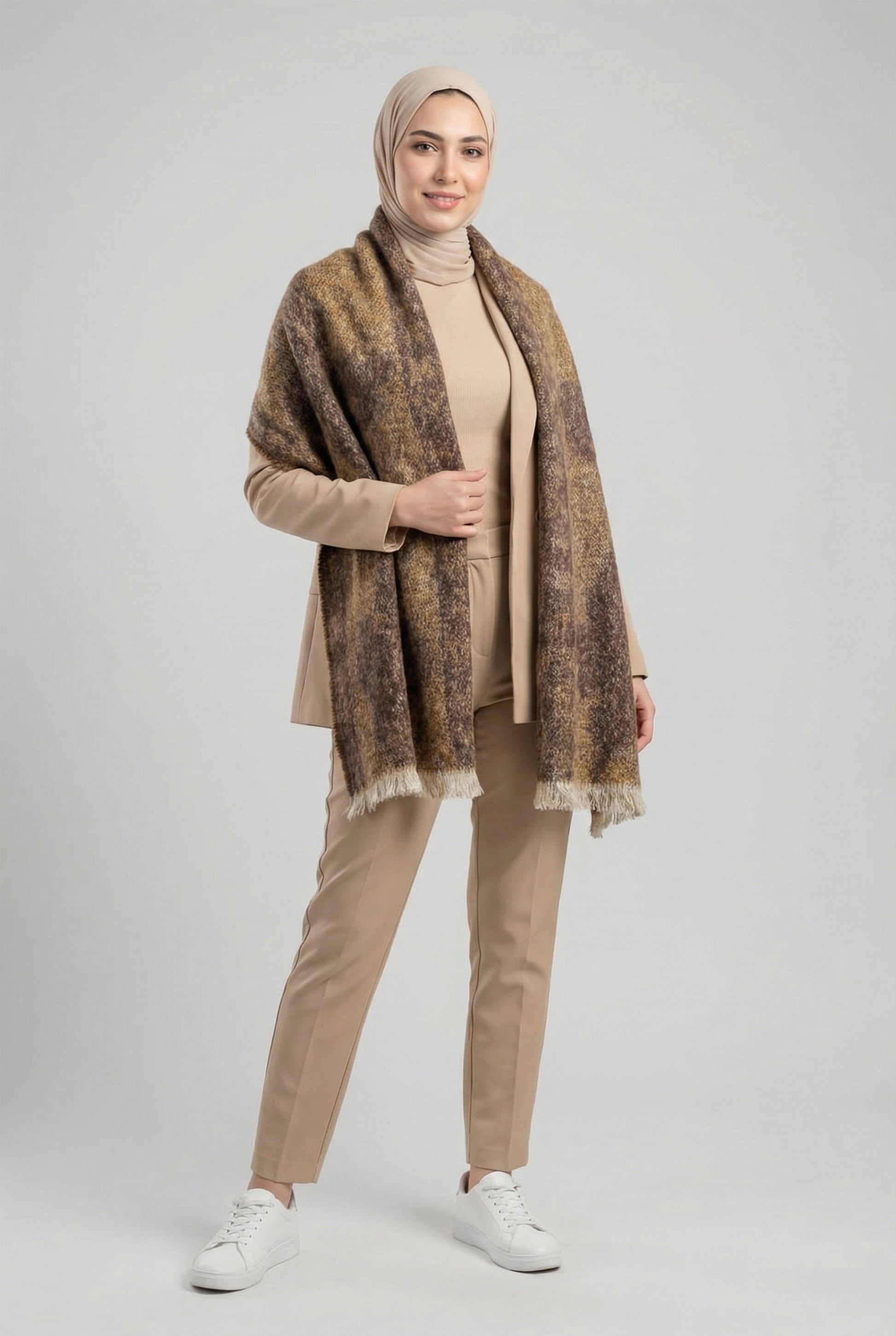 Woman wearing a patterned shawl, beige top, pants, and white shoes on a light gray background