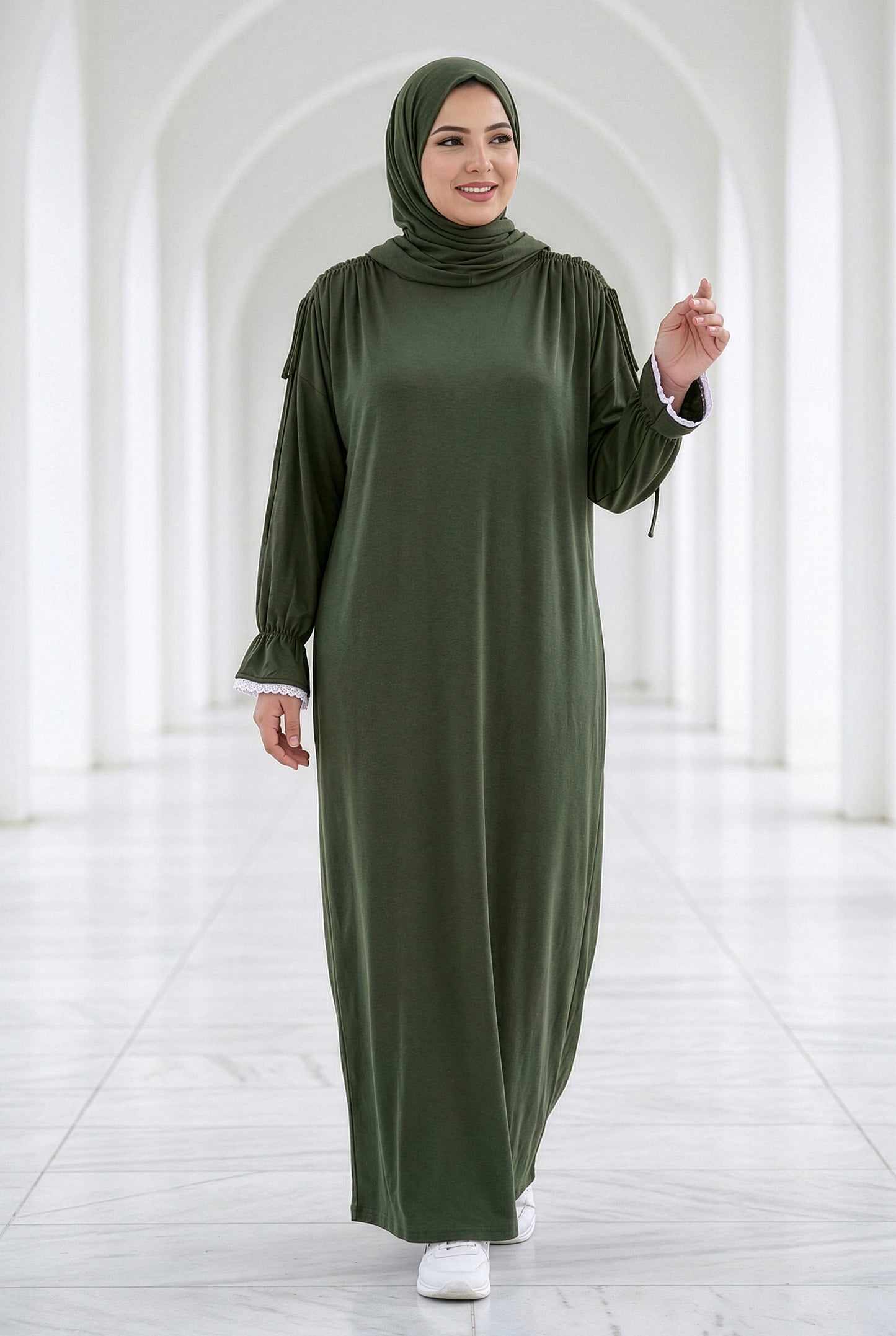 Woman wearing a dark olive Grace Isdal with white embroidered cuffs, modest full-length abaya photographed in a bright white arched hallway