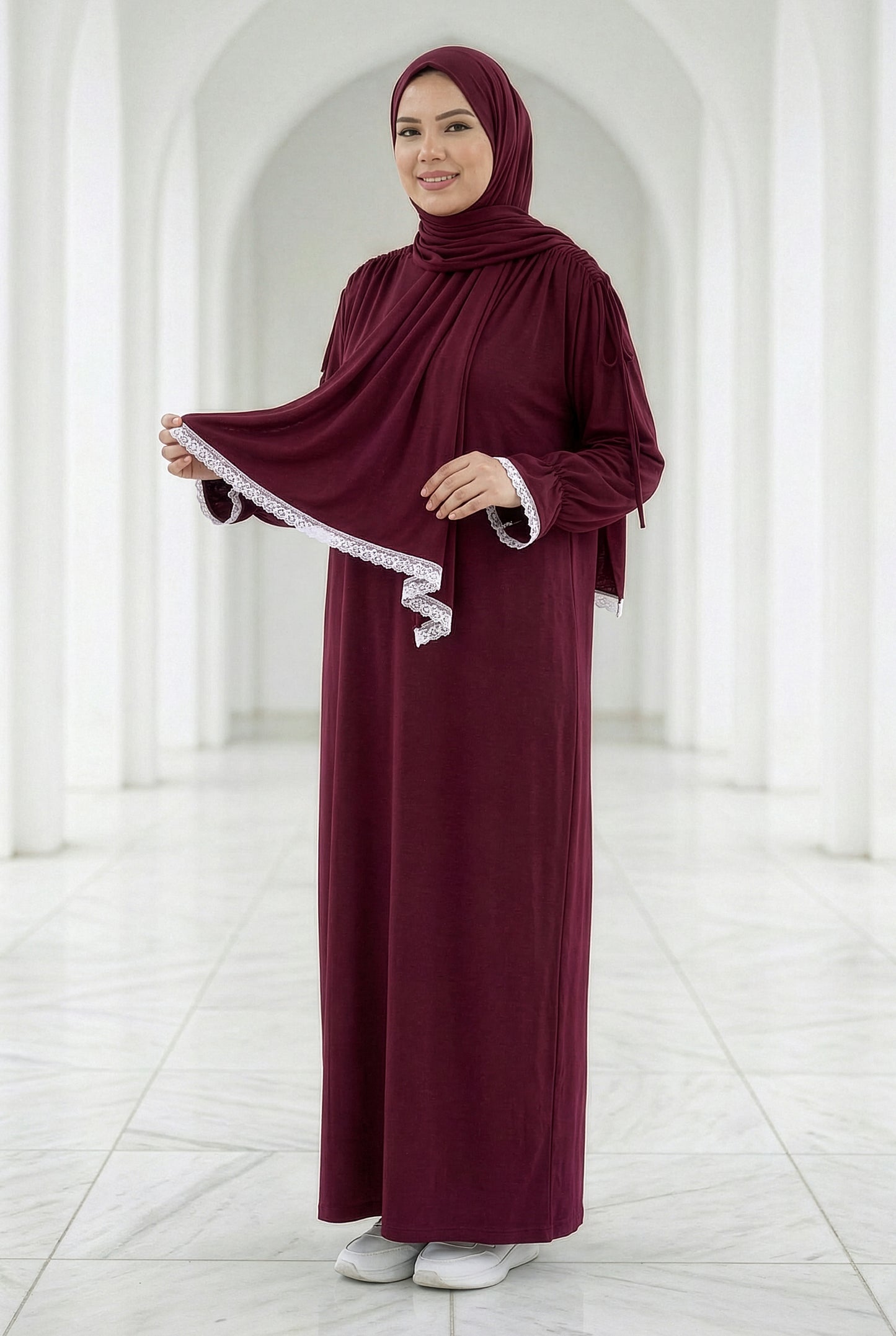 Woman wearing a deep burgundy Grace Isdal with white embroidered trim on the shawl and cuffs, modest flowing design in an elegant white archway setting