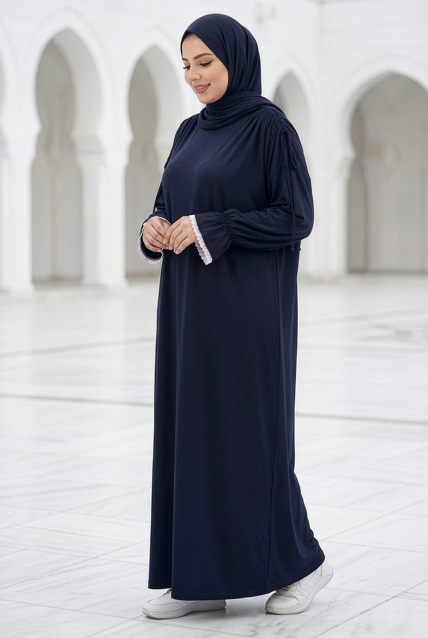 Woman wearing a deep navy Grace Isdal with white embroidered trim on the shawl and cuffs, modest flowing abaya photographed in an elegant white-arched courtyard