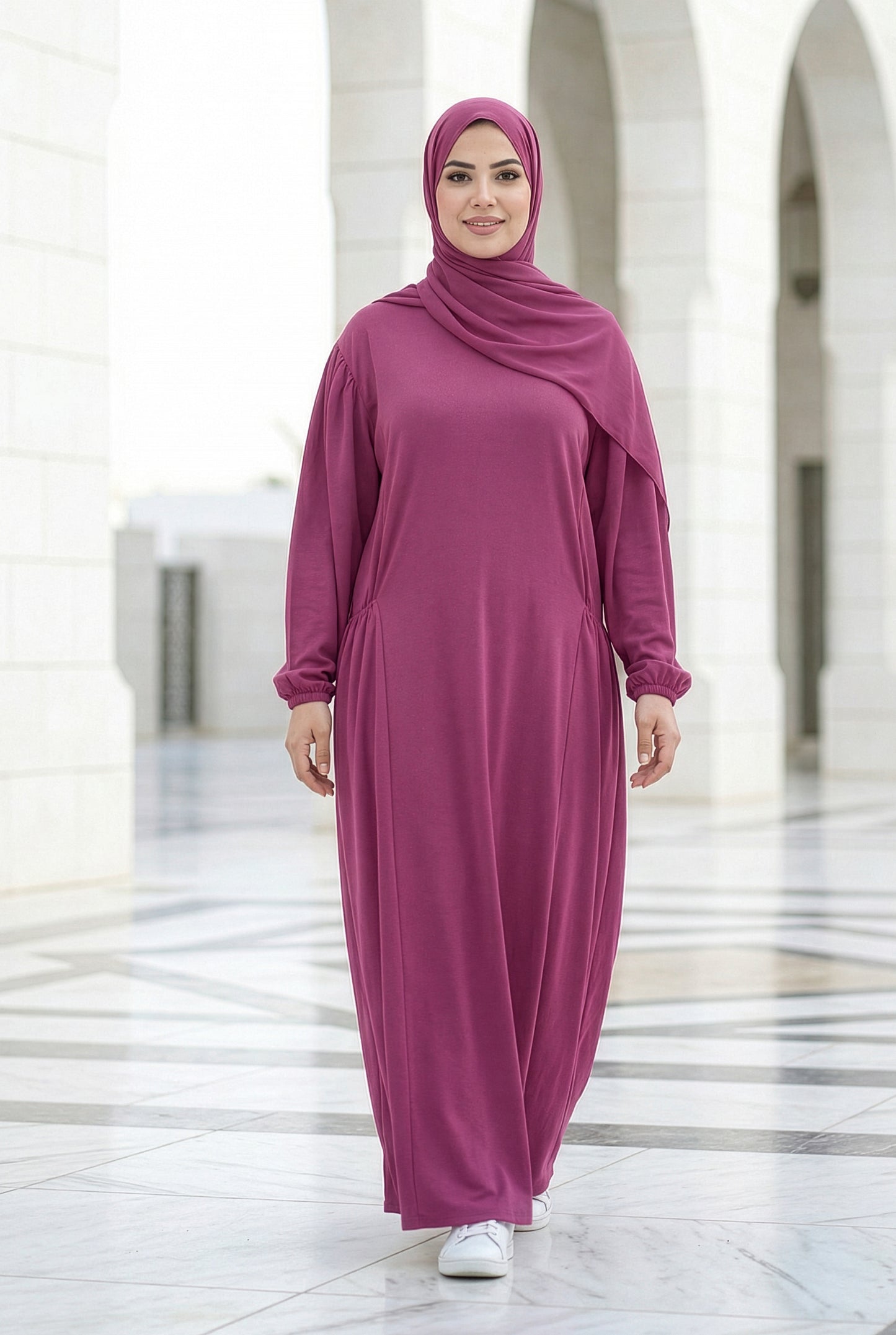 Woman wearing a dark fuchsia Royale Isdal, full-length modest abaya with a flowing relaxed fit, photographed in a bright outdoor marble courtyard.