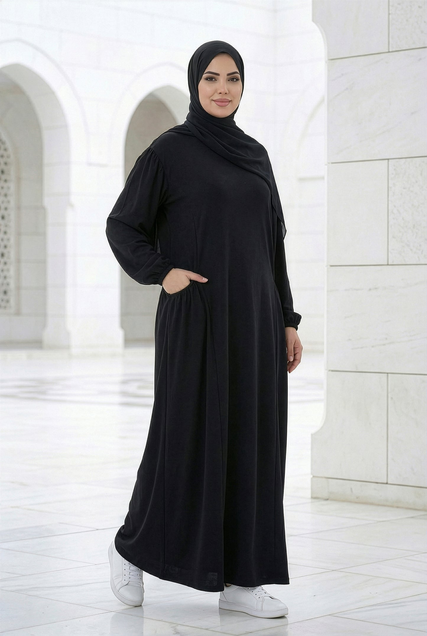 Woman wearing a black Royale Isdal with side pockets, modest full-length abaya photographed in a bright marble courtyard