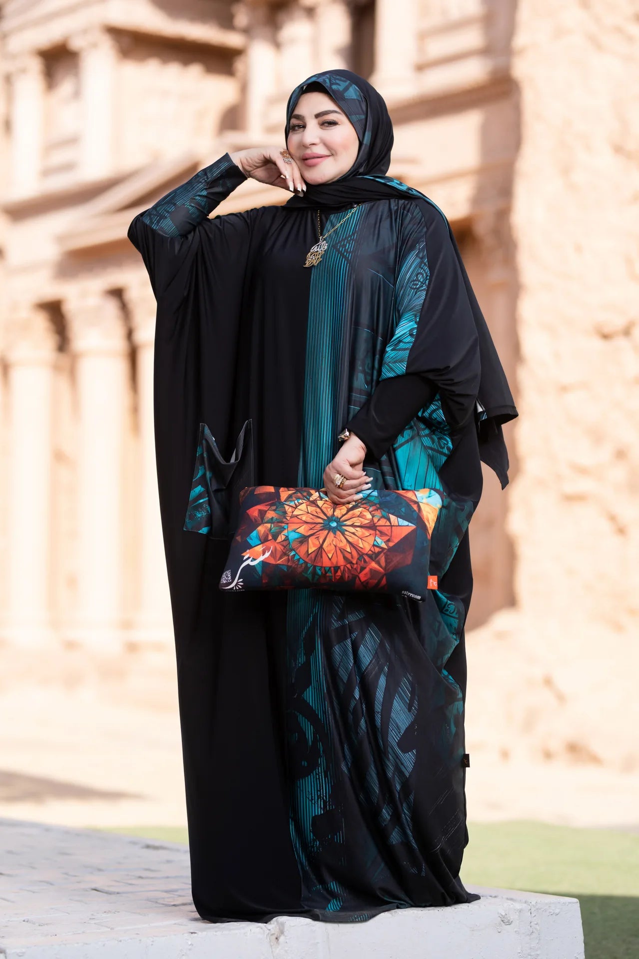 Woman in a black and teal abaya holding a colorful clutch against a beige wall.
