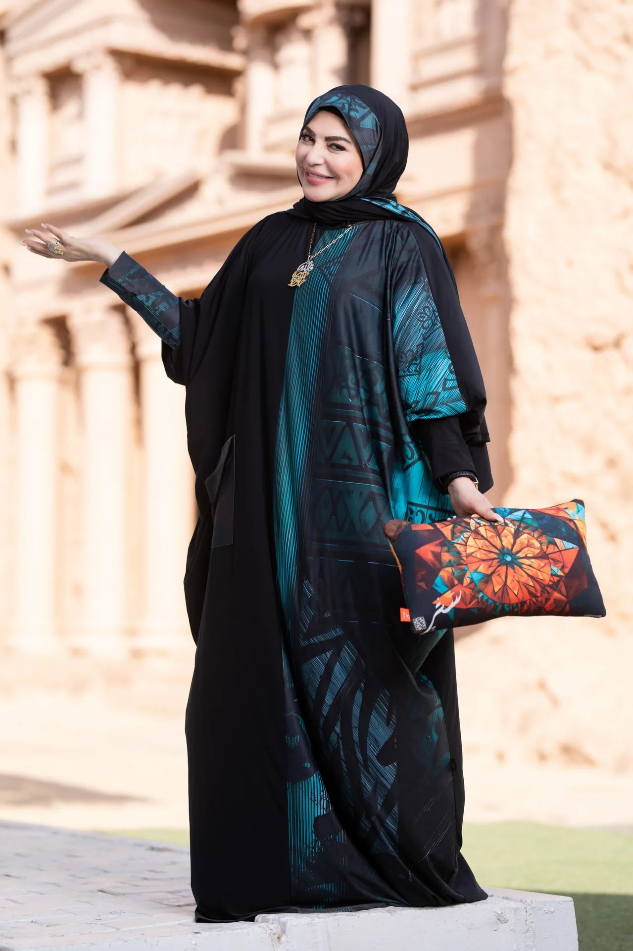 Woman in a black and teal abaya holding a colorful pillow outdoors.