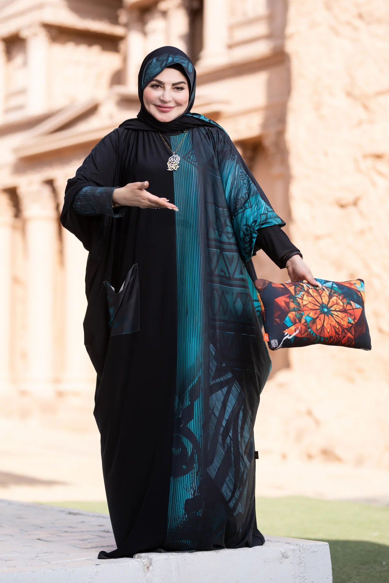 Woman in black abaya with teal patterned scarf holding a colorful pillow, standing in front of a stone wall.