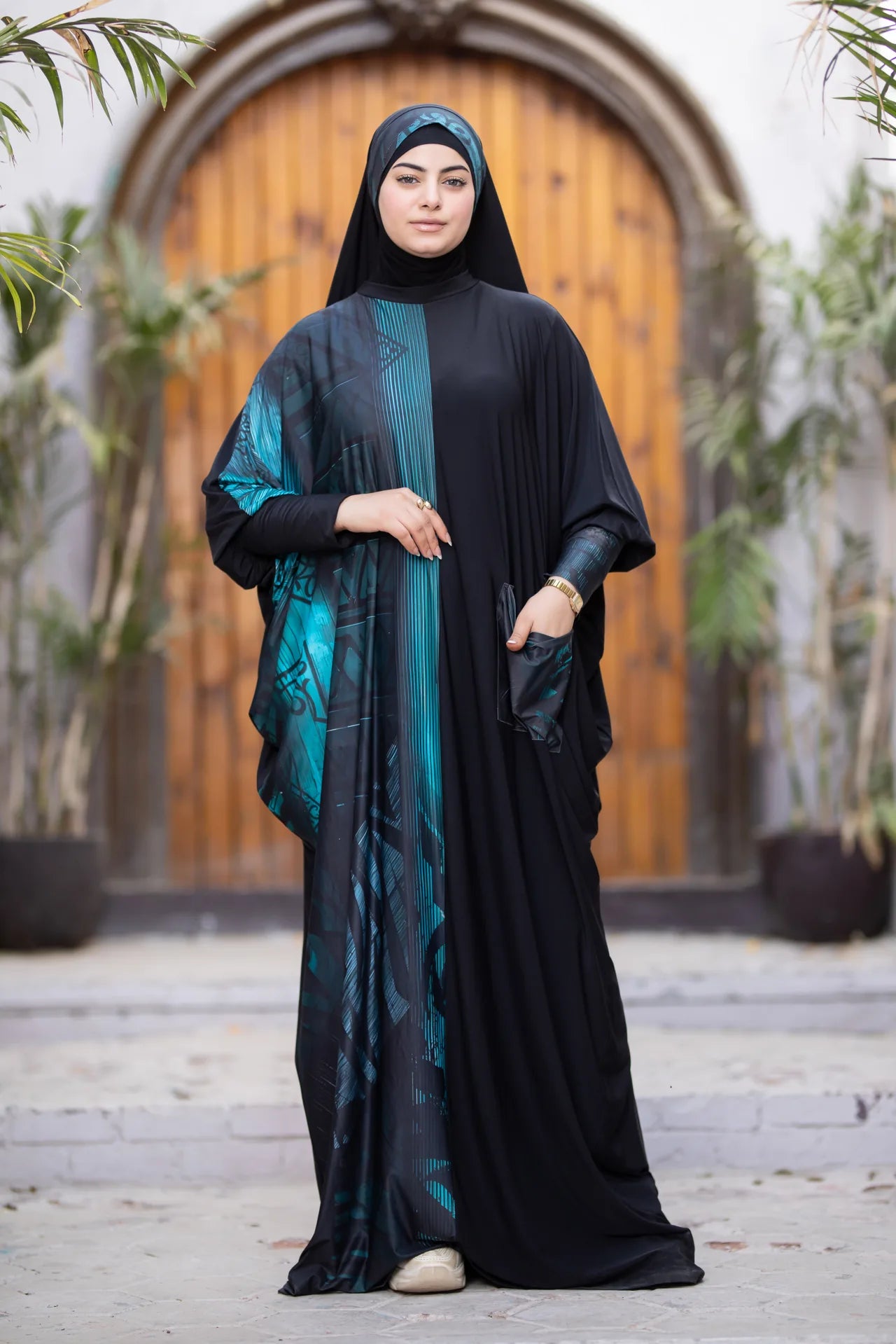 Woman wearing a black and teal abaya in front of a wooden door.