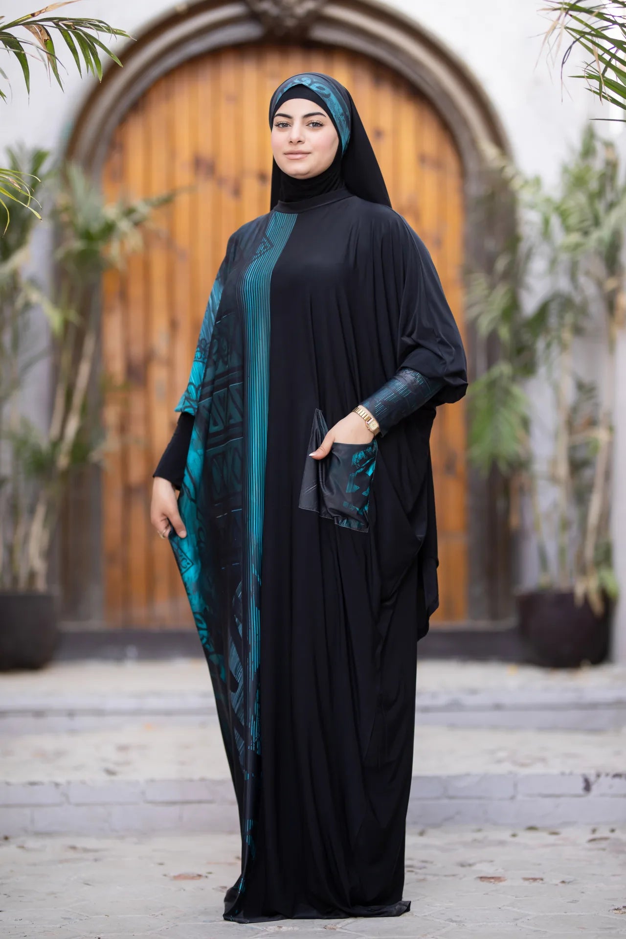 Woman in a black abaya with green accents standing in front of a wooden door.