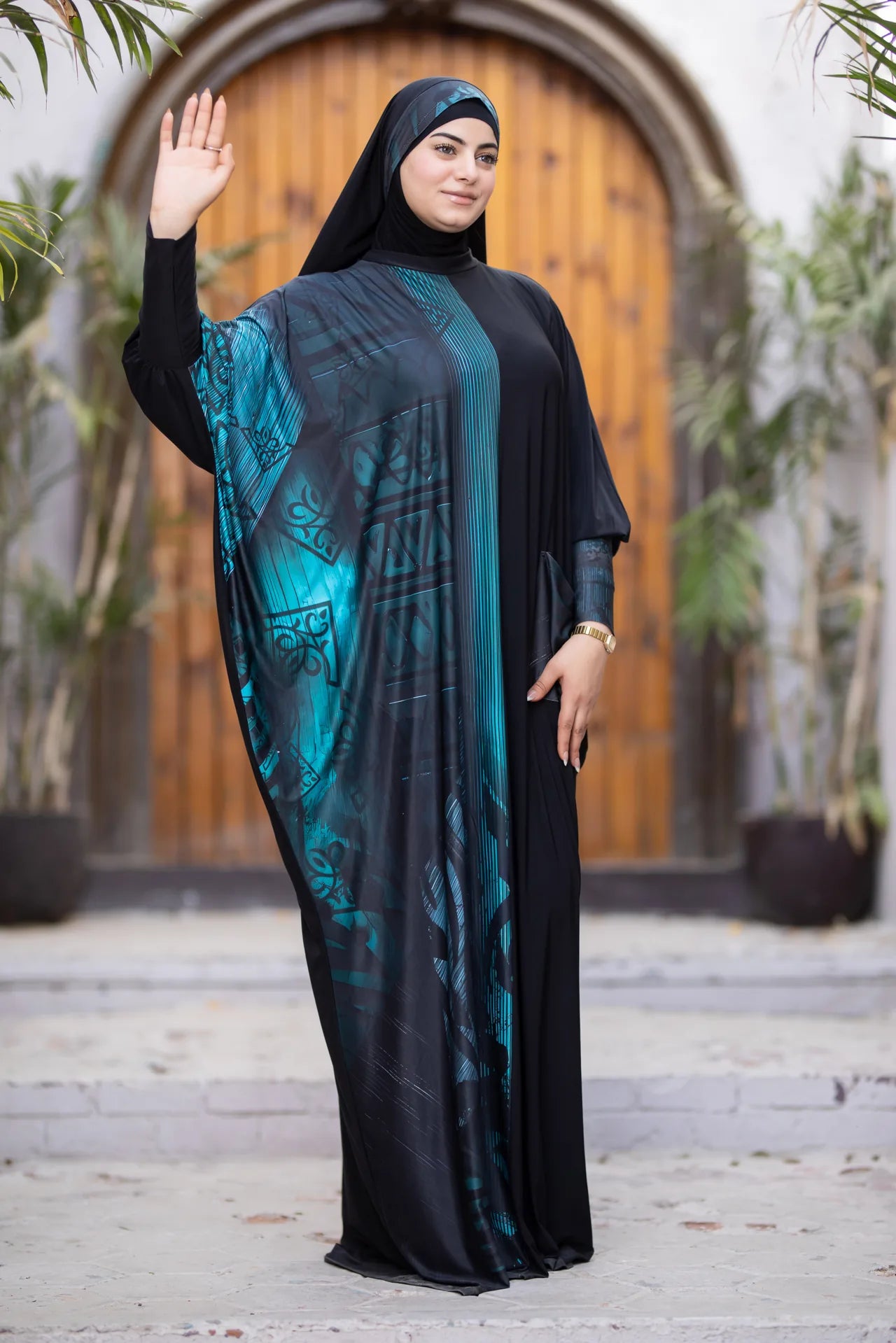 Woman wearing a black and teal abaya in front of a wooden door.
