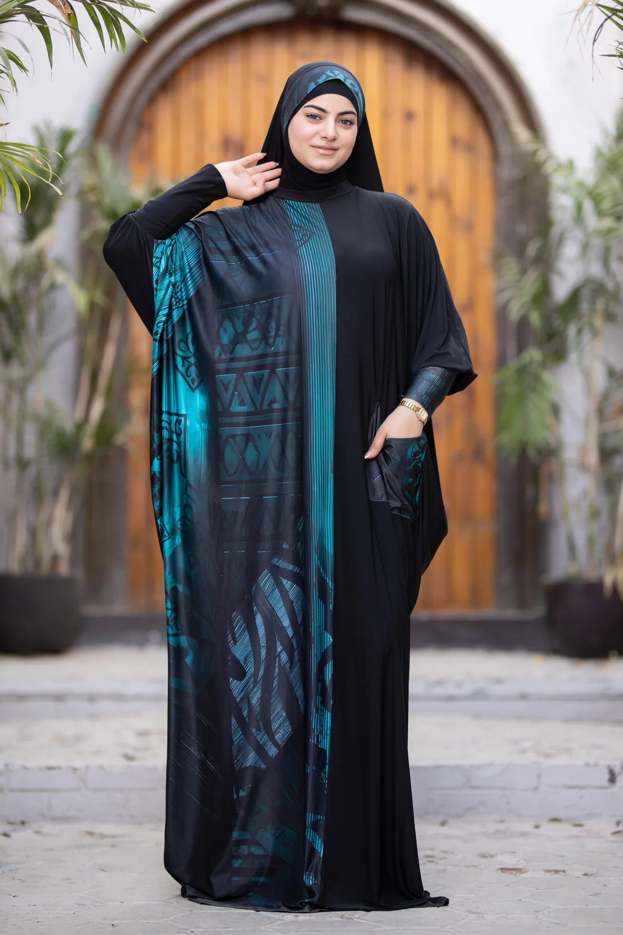 Woman wearing a black abaya with a teal pattern in front of a wooden door.
