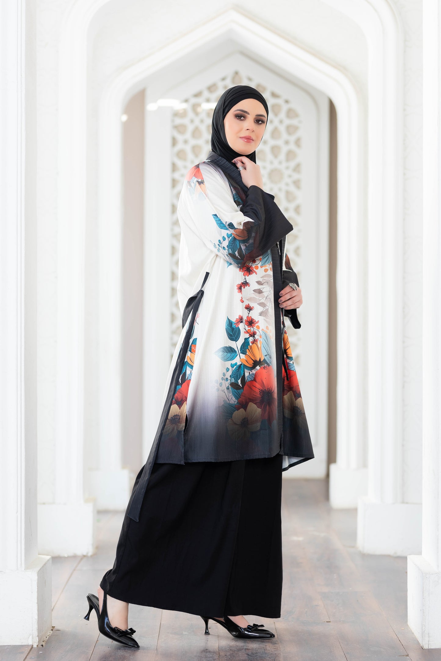 Vibrant floral printed long kimono robe with black base, worn over long black dress by a woman in hijab, standing in a traditional architecture frame