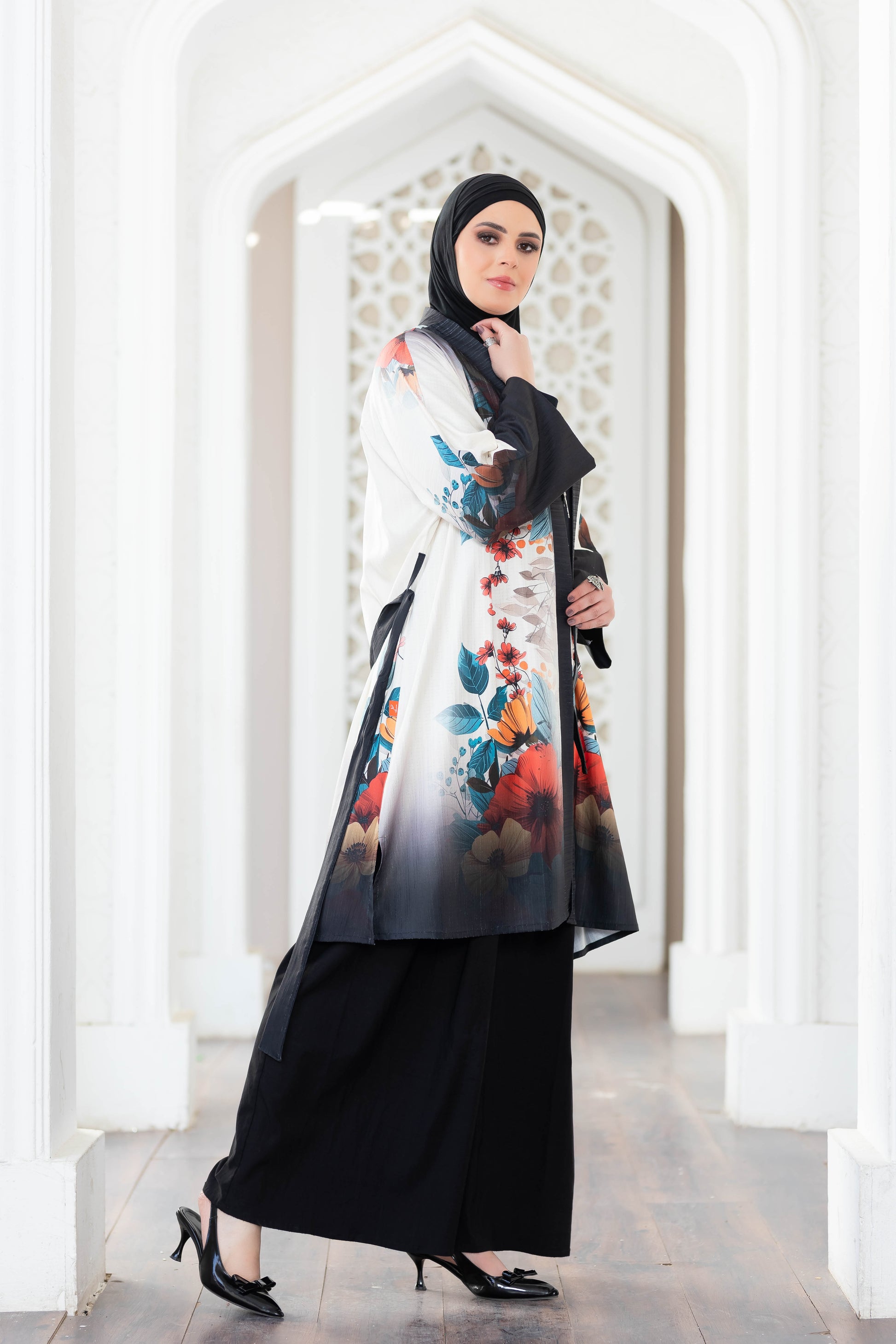 Vibrant floral printed long kimono robe with black base, worn over long black dress by a woman in hijab, standing in a traditional architecture frame