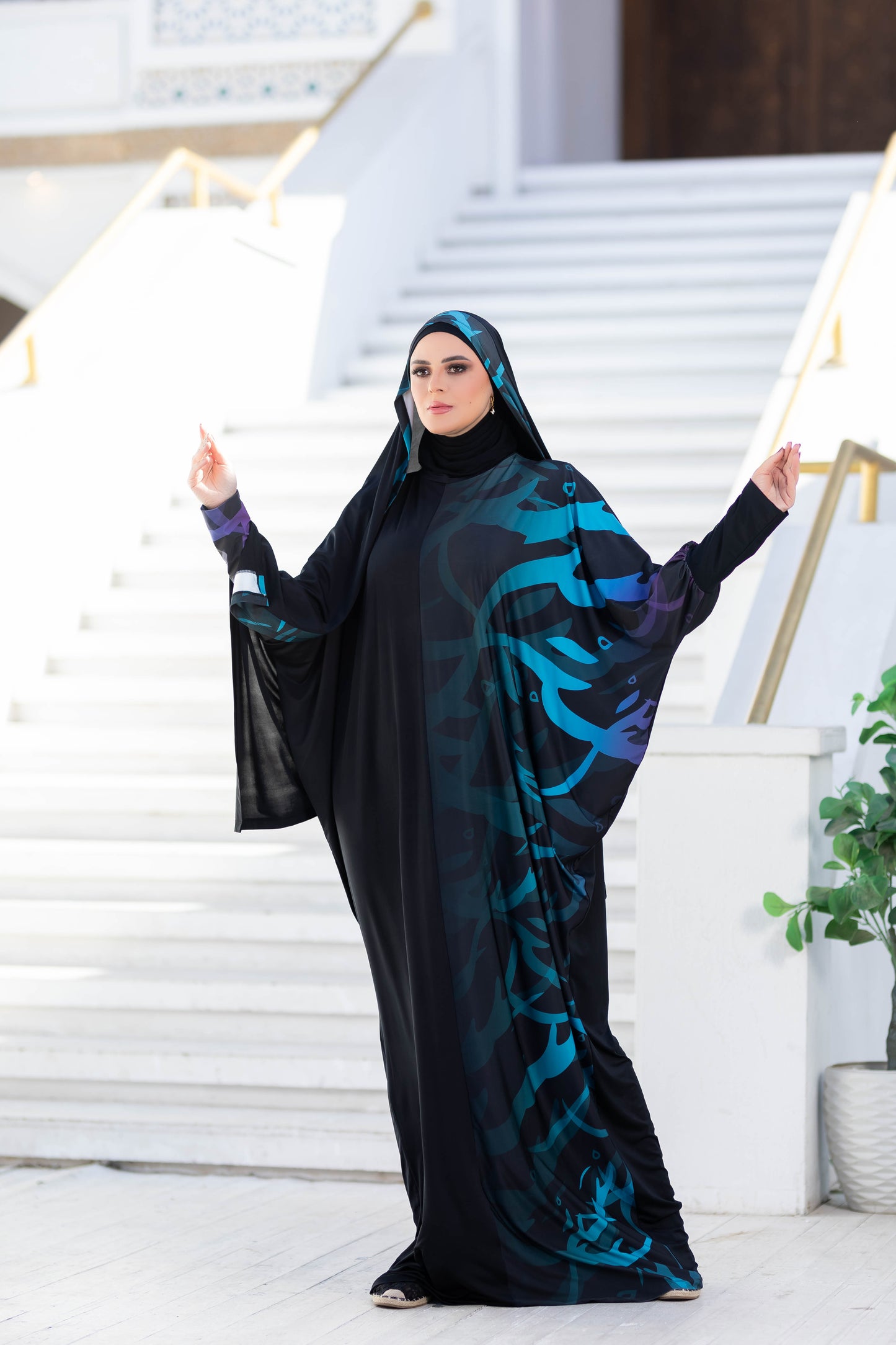 Elegant modest woman in traditional Islamic attire, wearing a black and teal printed long dress with a headscarf, standing on stairs against a light background.
