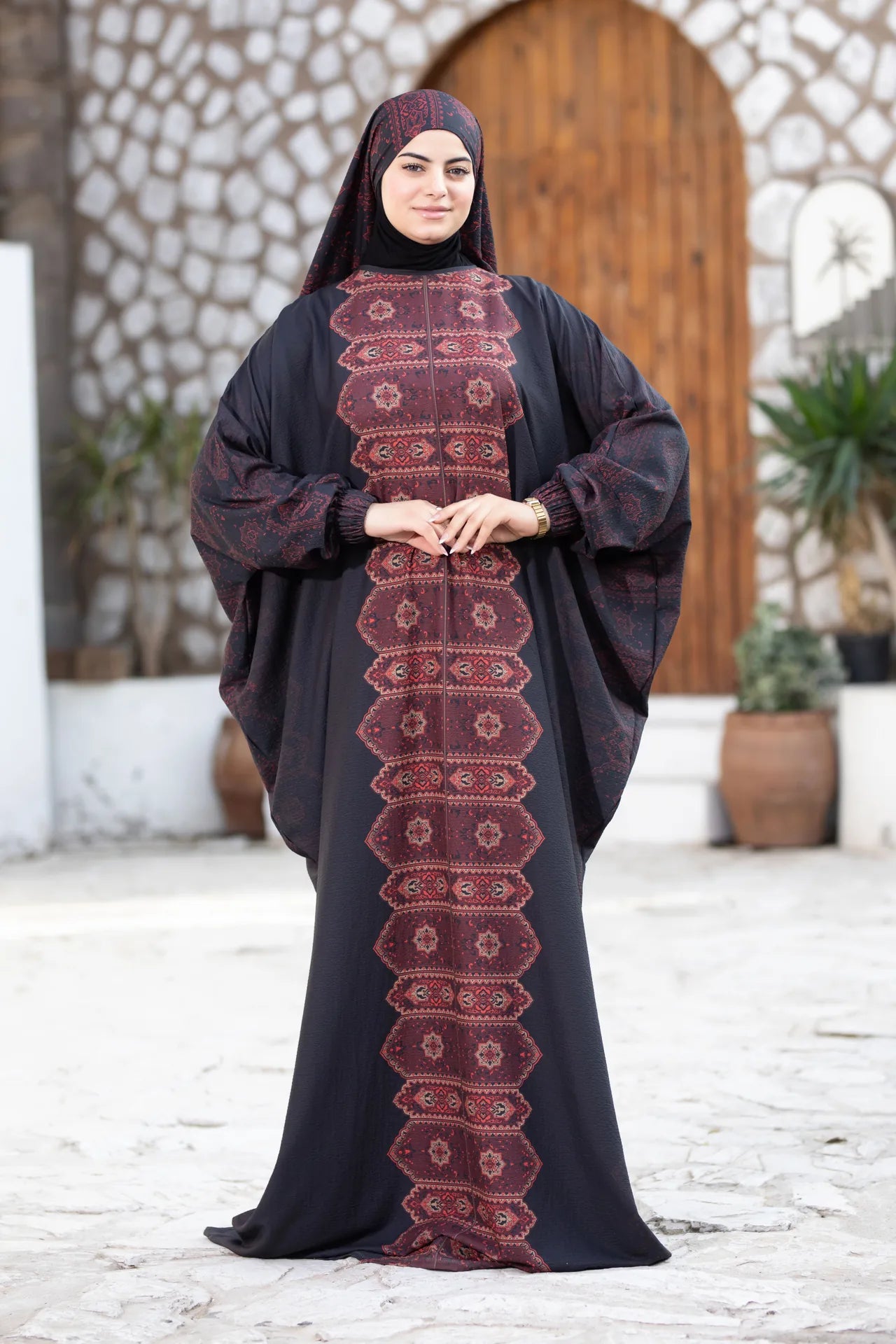 Woman wearing a dark abaya with intricate red patterns in an outdoor setting.