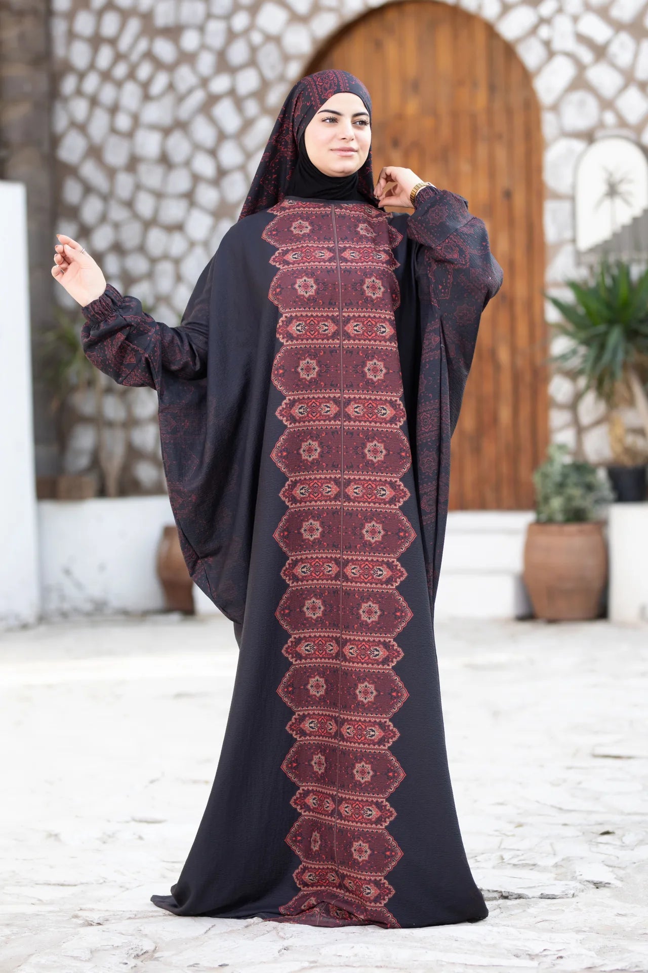 Woman wearing a black abaya with red patterns in an outdoor setting.