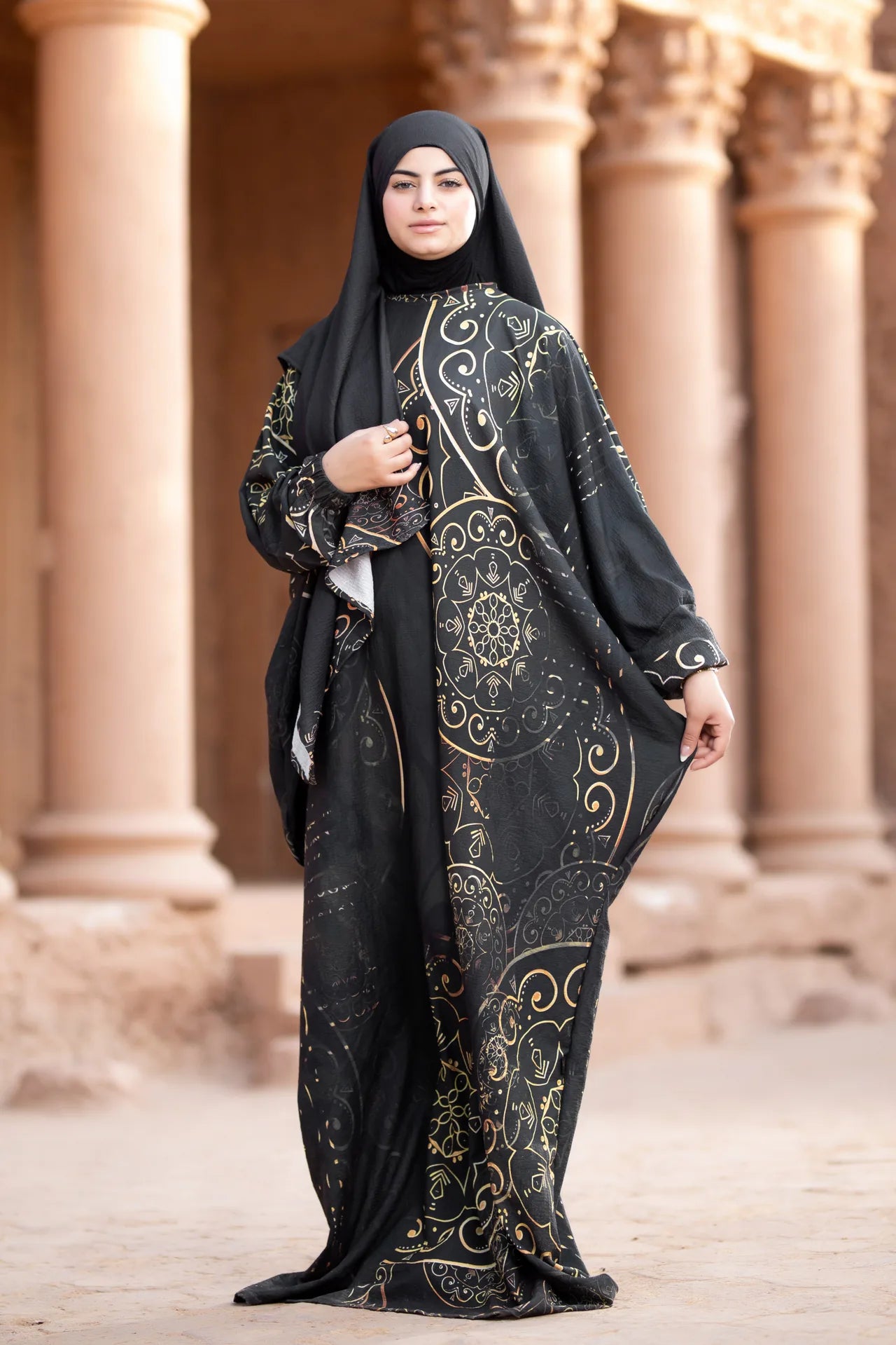 Woman wearing a black abaya with gold embroidery in front of classical architecture.