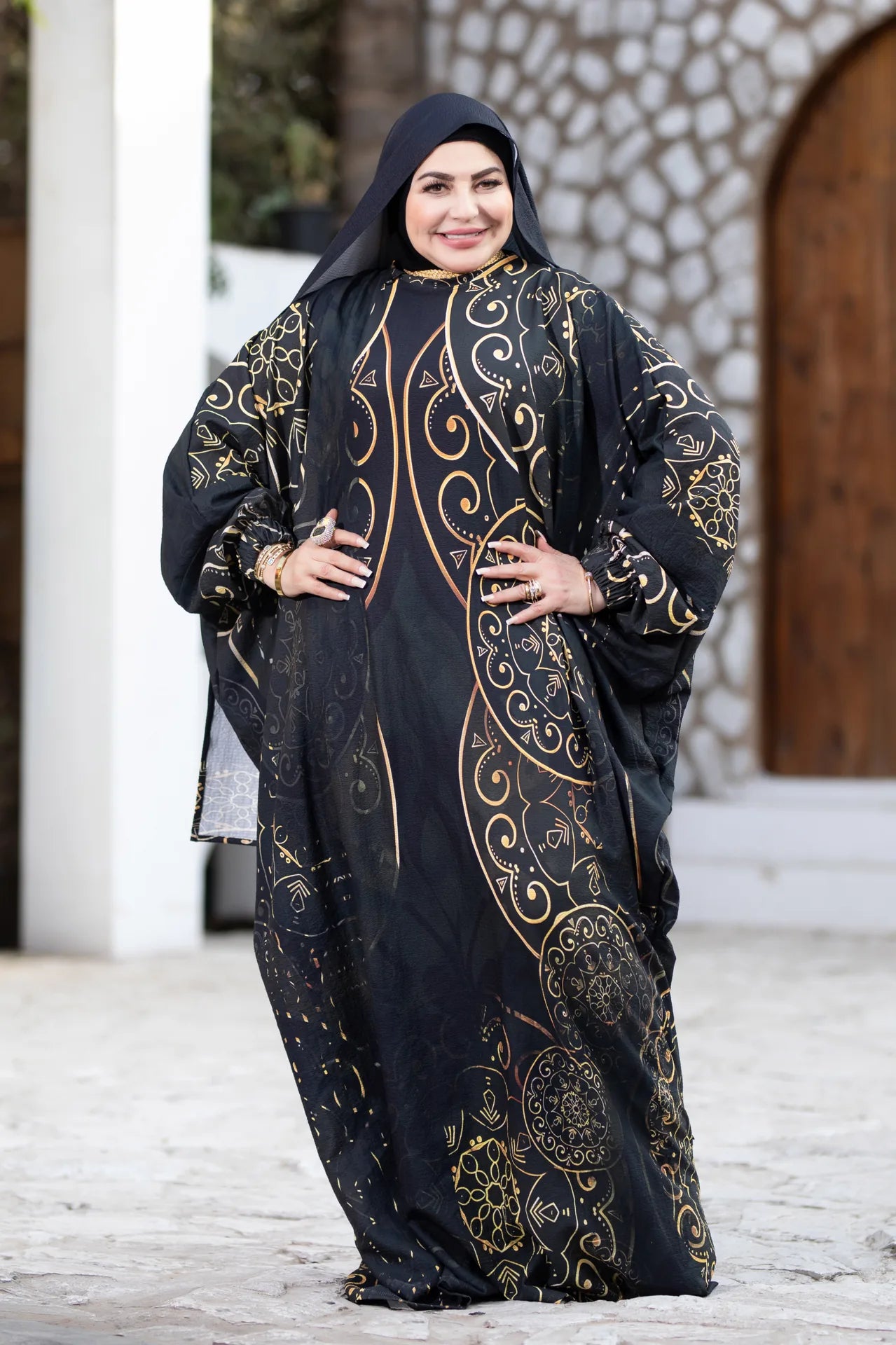 Woman wearing a black and gold embroidered abaya in front of a stone building.