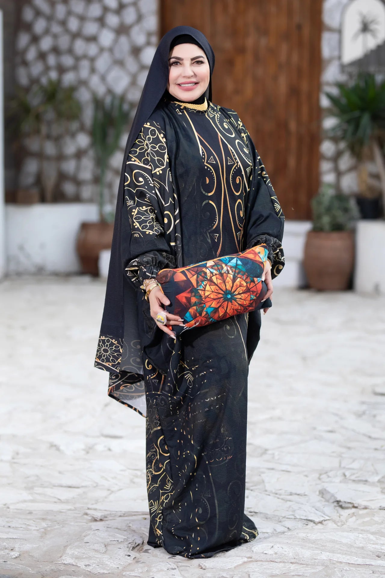 Woman in traditional black and gold outfit holding a colorful clutch against a stone wall background