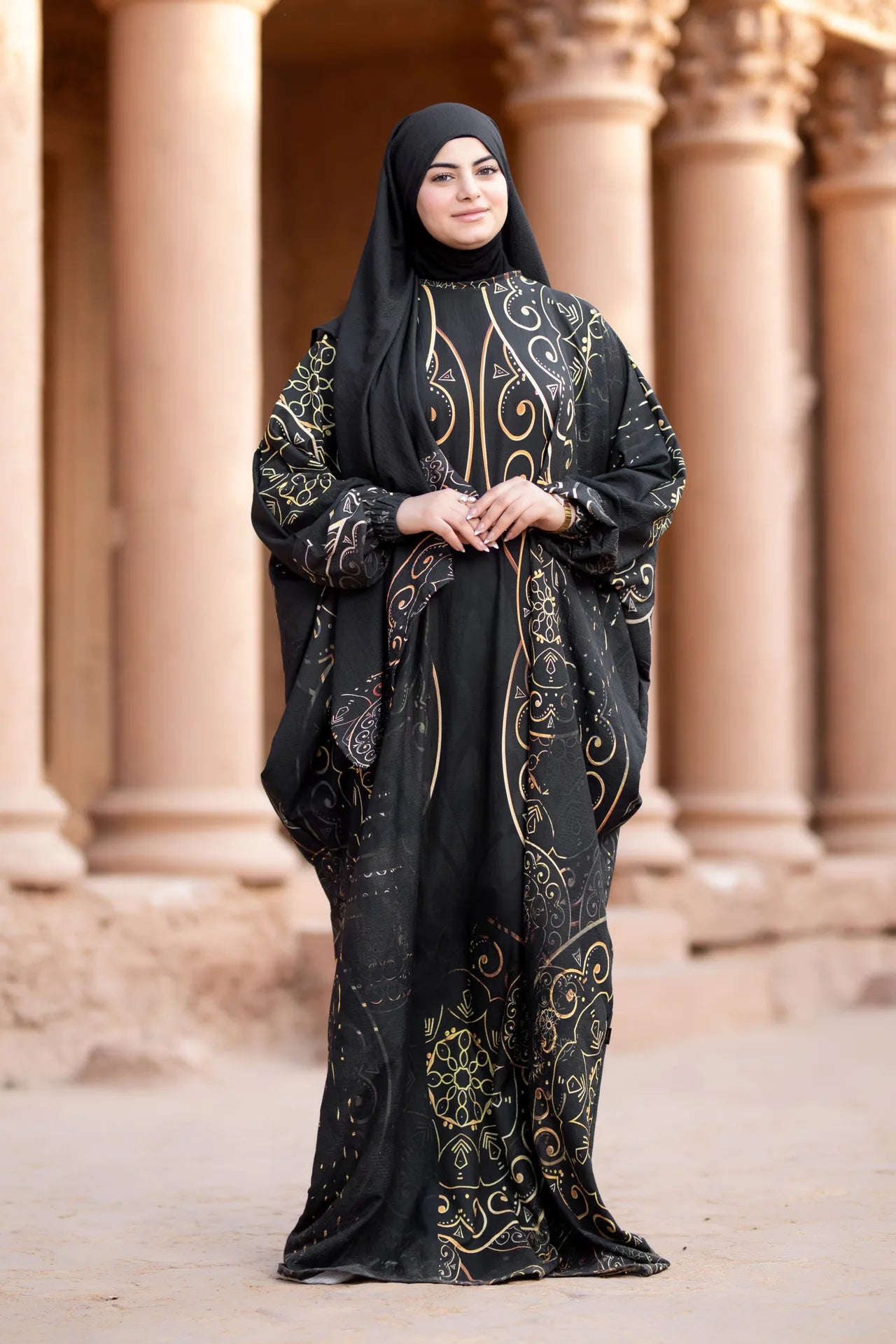 Woman wearing a black abaya with gold embroidery standing in front of stone columns.