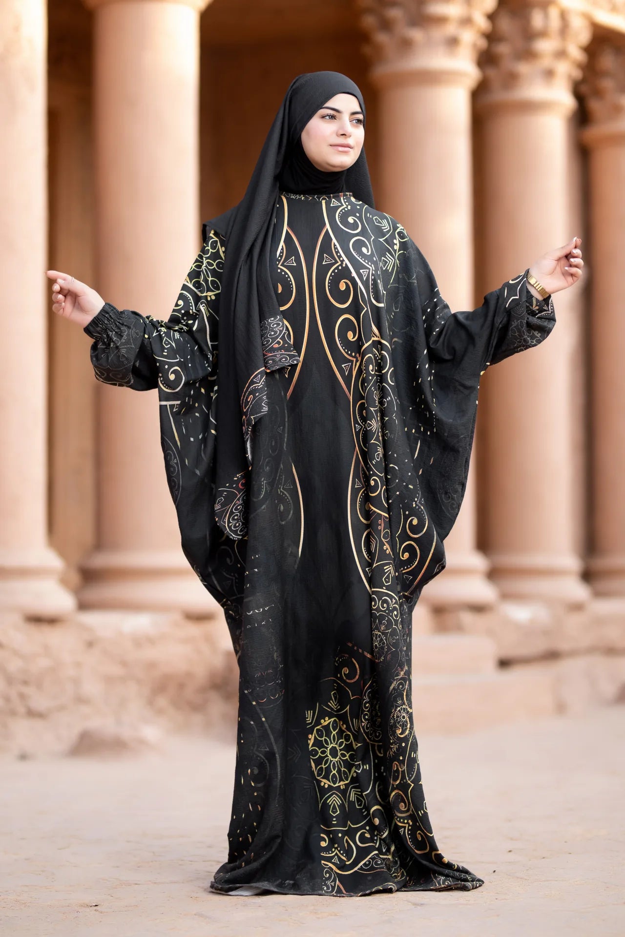 Woman wearing a black abaya with gold embroidery in front of classical architecture.