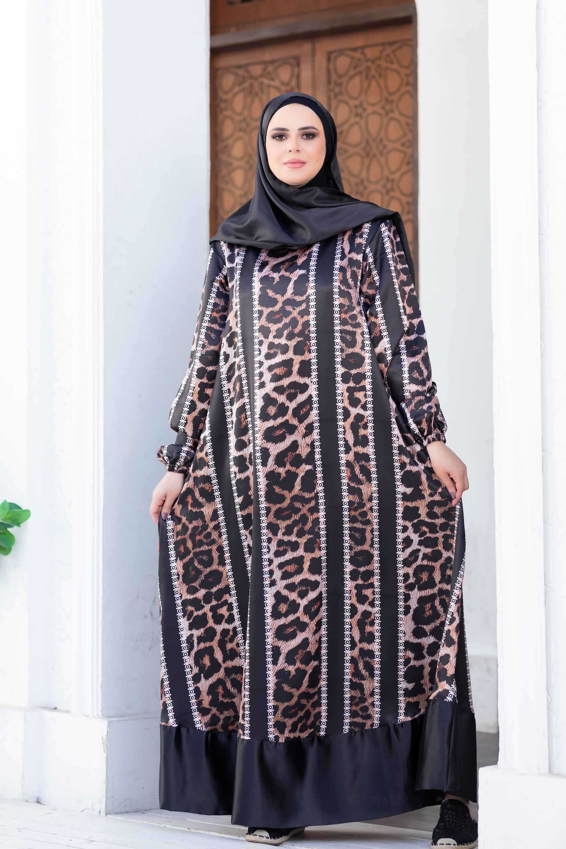 Elegant modest dress with leopard print pattern, black hijab, woman standing in front of traditional wooden door