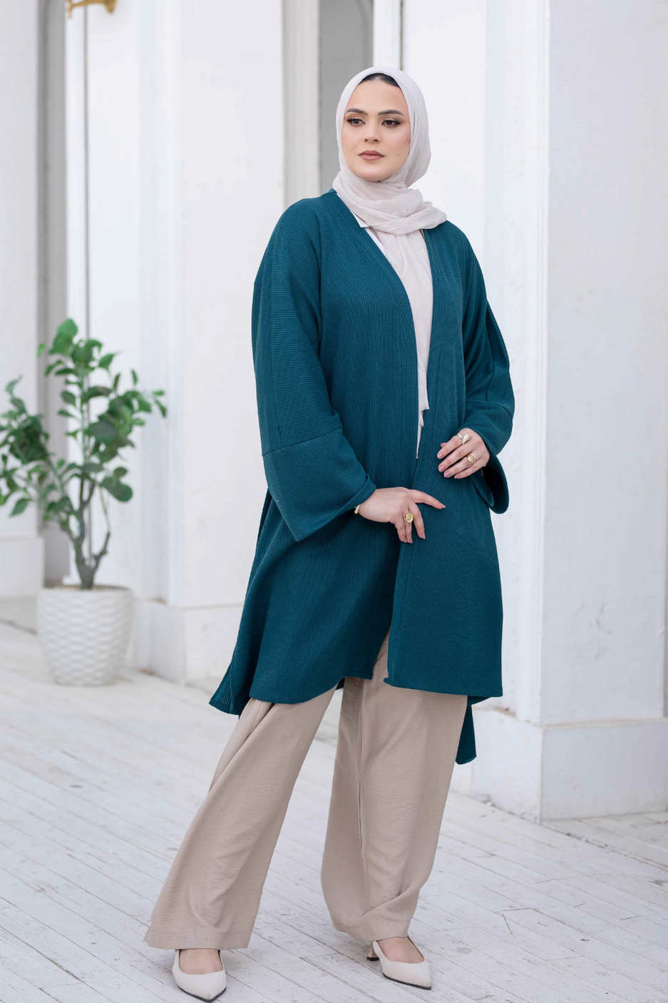 Elegant wool blend midi coat for women in teal blue, worn by a woman in a headscarf, standing against a white background with a plant in the foreground.