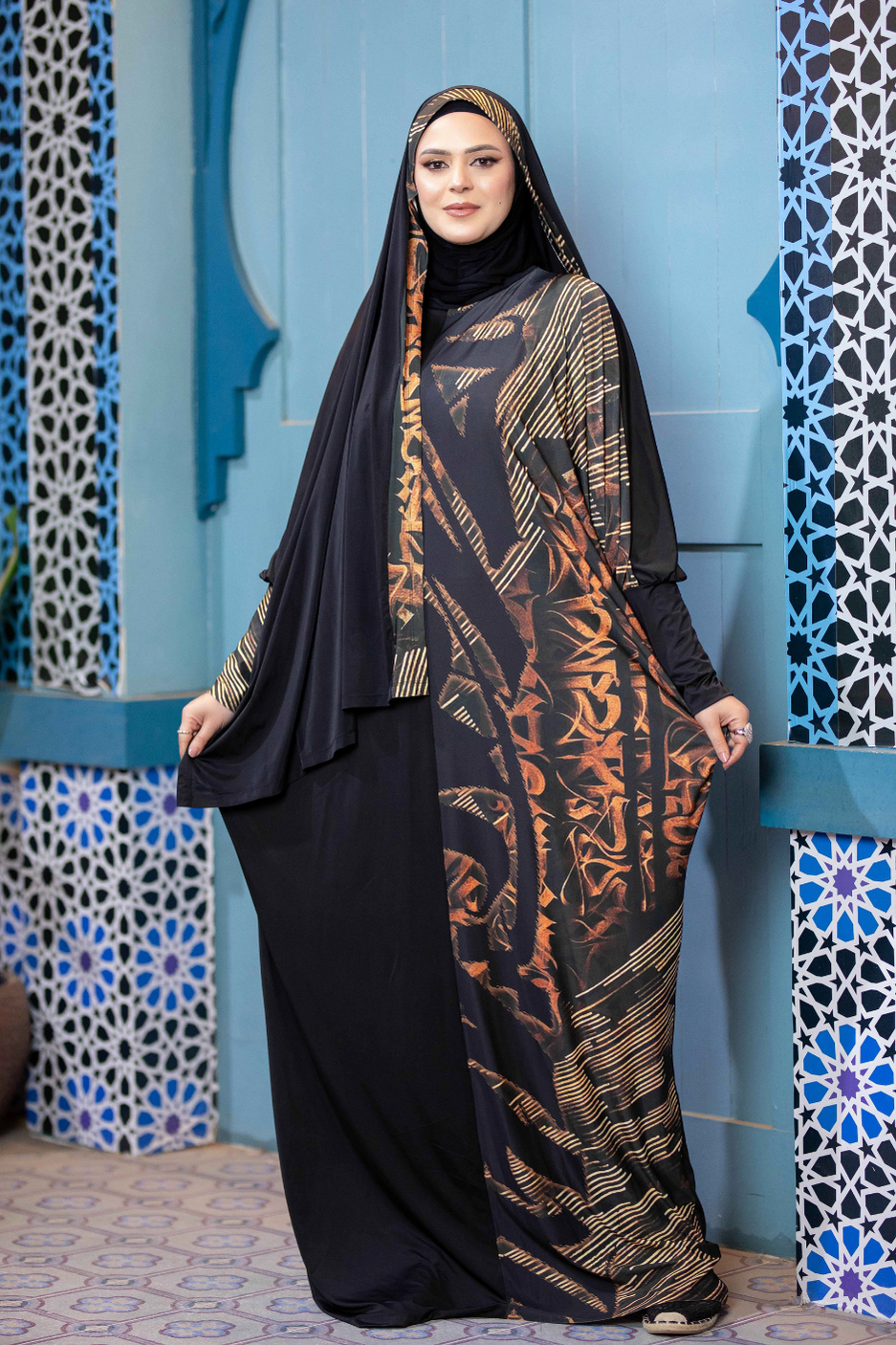 Elegant Islamic dress with intricate patterns and vibrant colors, featuring a long black robe and patterned scarf covering the model's head, set against a vibrant, tiled background.