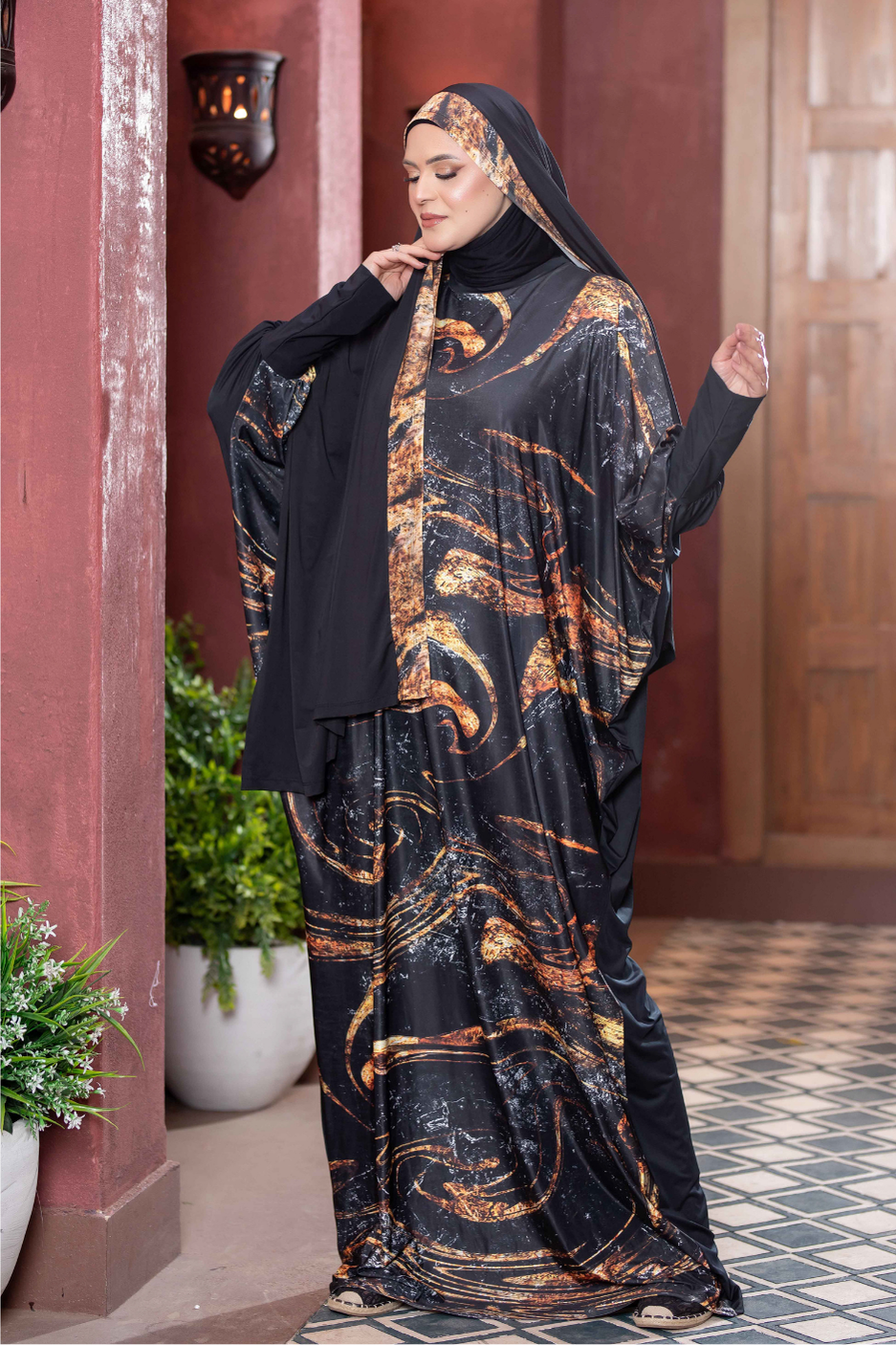 Elegant Islamic fashion: Black and orange patterned maxi dress with matching headpiece, worn by a woman standing against a traditional red wall backdrop.