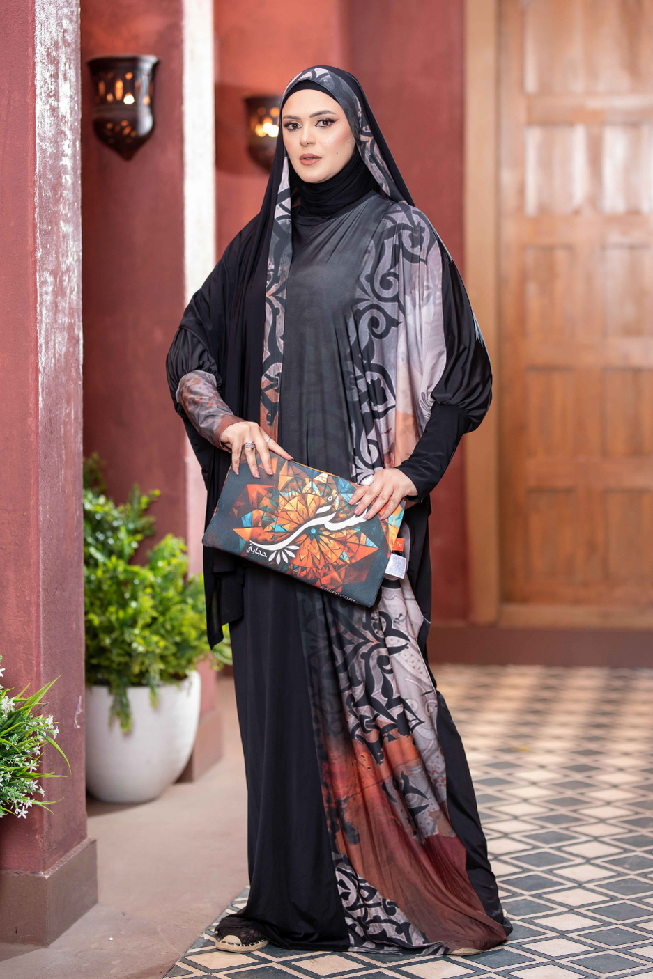 Modest Islamic fashion: Elegant woman in hijab holding patterned scarf, standing in front of ornate doorway.