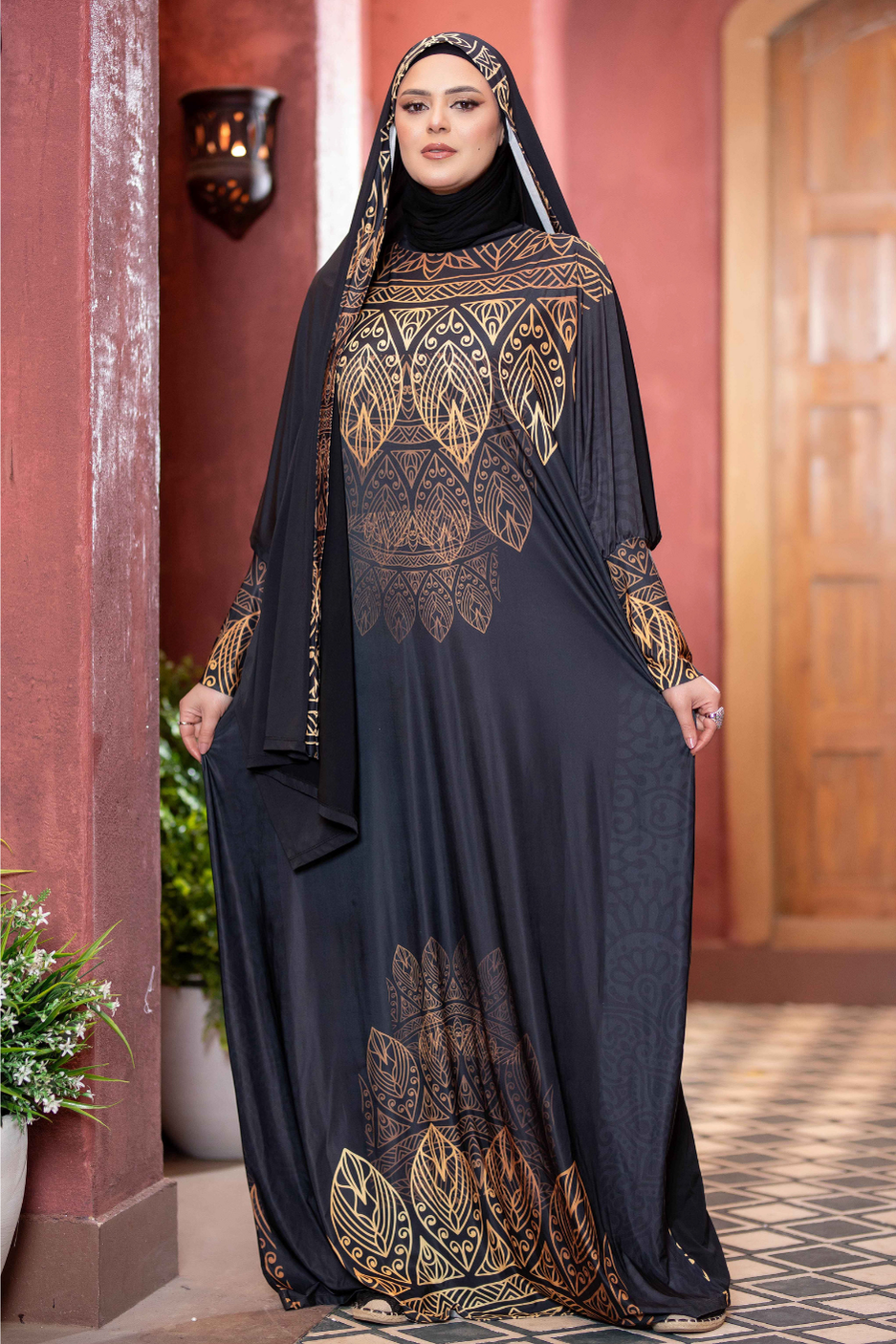 Elegant long black dress with ornate gold and black pattern, worn by woman with hijab in traditional Middle Eastern style, standing in archway.