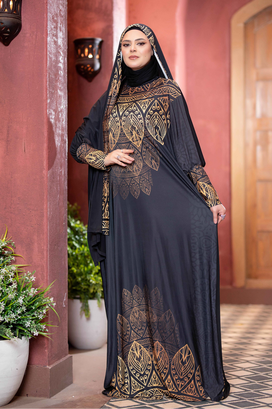 Elegant black and gold printed Islamic dress with intricate floral patterns, featured in the foreground of the image against a red wall background.