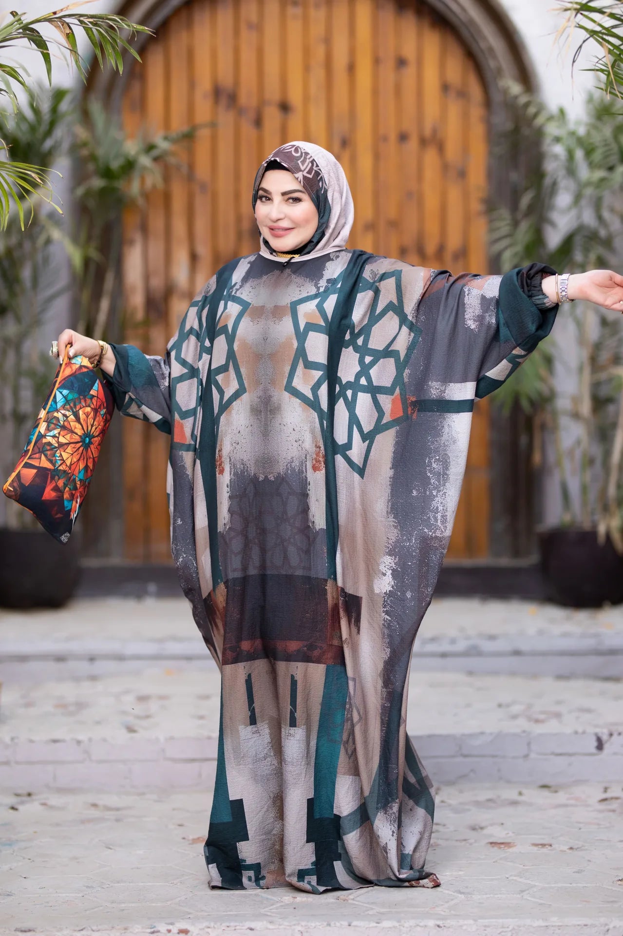 Woman in a patterned outfit holding a colorful bag outdoors with plants and a wooden door in the background.