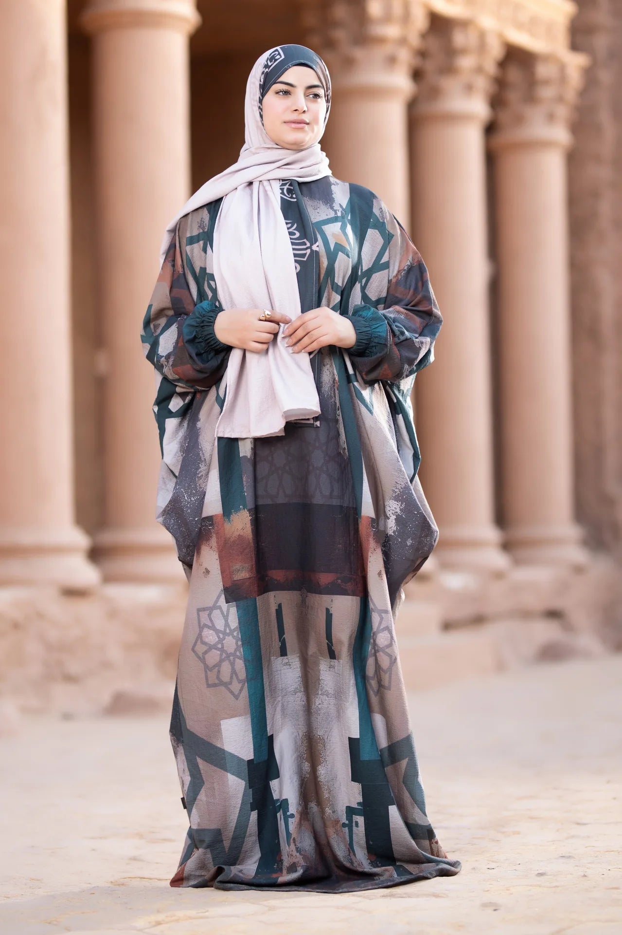 Woman wearing a patterned dress and hijab standing in front of architectural columns.